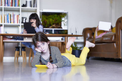 Kleines Mädchen mit Smartphone, das zu Hause auf dem Boden liegt, während ihre Schwestern im Hintergrund digitale Tablets benutzen - ERRF03205