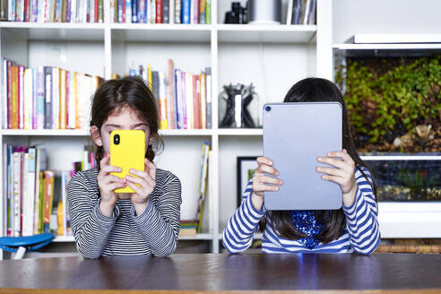 Two sisters sitting side by side at table in the living room using electronic devices - ERRF03207