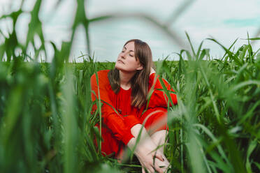 Portrait of woman wearing red dress sitting in a field with eyes closed - ERRF03229