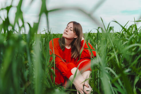 Portrait of woman wearing red dress sitting in a field with eyes closed - ERRF03229