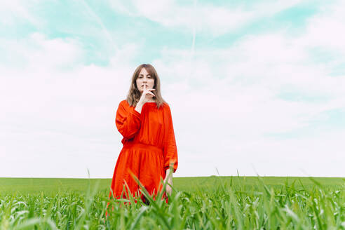Portrait of woman wearing red dress standing in a field with finger on mouth - ERRF03237