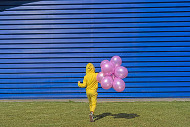 Back view of girl with pink balloons wearing yellow tracksuit walking in front of blue background - ERRF03242