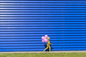 Kleines Mädchen mit rosa Luftballons und gelbem Trainingsanzug vor blauem Hintergrund - ERRF03244