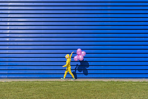 Kleines Mädchen mit Kopfhörern, das einen gelben Trainingsanzug trägt und vor einem blauen Hintergrund mit rosa Luftballons läuft - ERRF03245