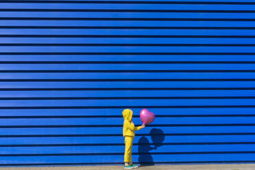 Little girl wearing yellow tracksuit standing with pink balloon in front of blue background - ERRF03249