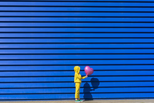 Kleines Mädchen im gelben Trainingsanzug steht mit rosa Luftballon vor blauem Hintergrund - ERRF03249