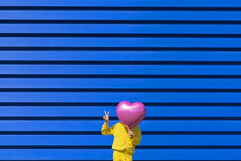 Little girl standing in front of blue background hiding behind pink balloon and showing victory sign stock photo