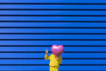 Little girl standing in front of blue background hiding behind pink balloon and showing victory sign - ERRF03250