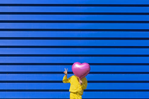 Kleines Mädchen steht vor blauem Hintergrund versteckt hinter rosa Ballon und zeigt Sieg Zeichen - ERRF03250