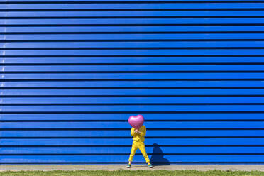 Little girl wearing yellow tracksuit standing in front of blue background hiding behind pink balloon - ERRF03251