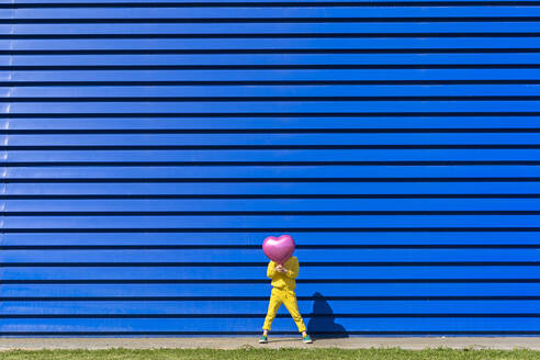 Kleines Mädchen im gelben Trainingsanzug vor blauem Hintergrund versteckt hinter rosa Luftballon - ERRF03251