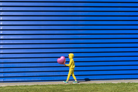 Kleines Mädchen im gelben Trainingsanzug geht mit rosa Luftballon vor blauem Hintergrund, lizenzfreies Stockfoto