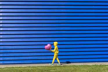 Kleines Mädchen im gelben Trainingsanzug geht mit rosa Luftballon vor blauem Hintergrund - ERRF03252