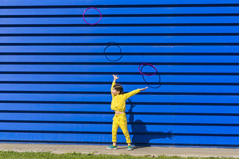 Kleines Mädchen im gelben Trainingsanzug wirft Gymnastikringe in die Luft vor blauem Hintergrund - ERRF03253