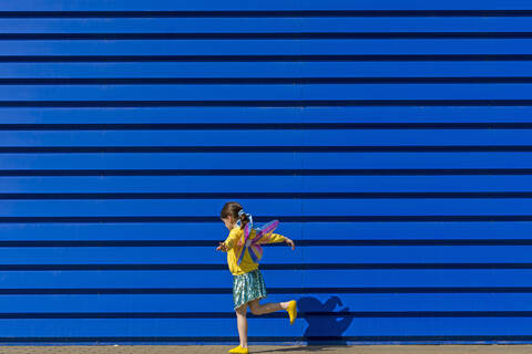 Kleines Mädchen mit bunten Schmetterlingsflügeln balanciert auf einem Bein vor blauem Hintergrund, lizenzfreies Stockfoto
