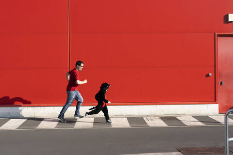 Mann läuft zusammen mit seinem kleinen Mädchen in schwarzem Kostüm über einen Zebrastreifen, lizenzfreies Stockfoto
