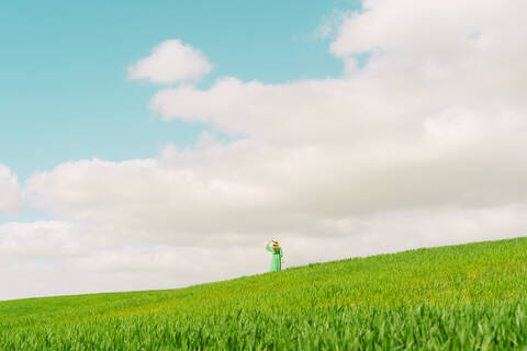 Rückenansicht einer Frau in grünem Kleid und Sommerhut, die auf einem Feld steht und in die Ferne schaut, lizenzfreies Stockfoto