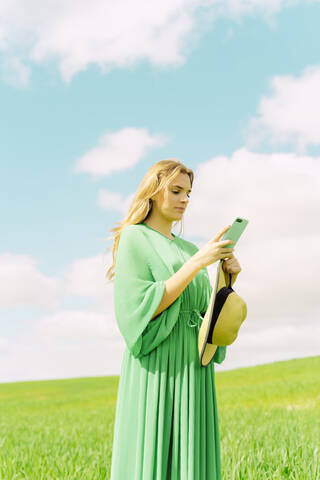 Junge Frau in grünem Kleid steht auf einem Feld und benutzt ein Smartphone, lizenzfreies Stockfoto