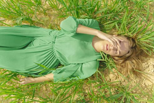 Blonde junge Frau in grünem Kleid liegt mit geschlossenen Augen auf einem Feld - ERRF03301
