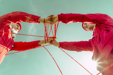 Crop view of young couple dressed in red performing with red string against sky - ERRF03382