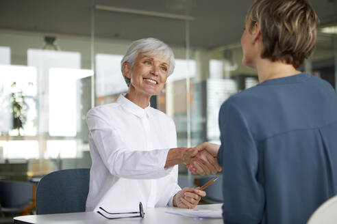 Two businesswomen shaking hands at desk in office - RBF07424