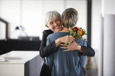 Glückliche ältere Geschäftsfrau mit Blumenstrauß, die einen Kollegen im Büro umarmt - RBF07427