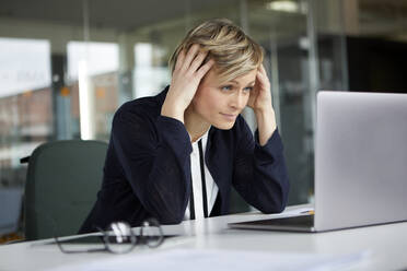 Businesswoman in office with head in hands using laptop - RBF07436