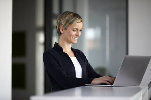 Lächelnde Geschäftsfrau mit Laptop im Büro - RBF07452