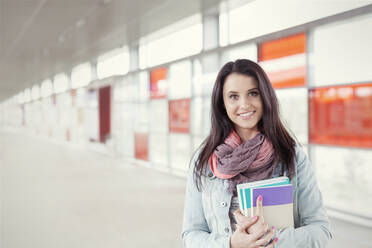 Portrait confident young woman with books standing in urban corridor - CAIF25785