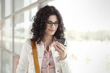 Young woman using smart phone at window - CAIF25786