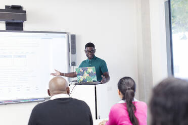 Männlicher Community-College-Lehrer, der an einem Laptop und einer Projektionsfläche im Klassenzimmer unterrichtet - CAIF25831