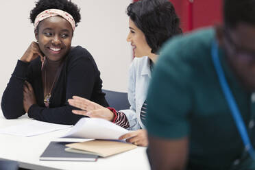 Smiling female community college students discussing paperwork in classroom - CAIF25868