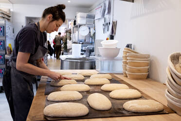 Woman wearing apron standing in an artisan bakery, shaping sourdough loaves for baking. - MINF14520