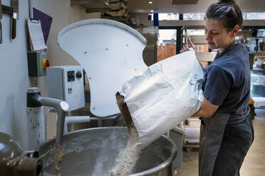 Eine Frau mit Schürze steht in einer handwerklichen Bäckerei und schüttet Mehl in einen industriellen Mischer. - MINF14527