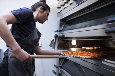 Eine Frau mit Schürze steht in einer handwerklichen Bäckerei und schiebt eine Pizza in den Ofen. - MINF14528