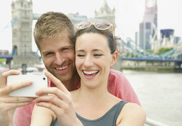 Happy couple taking selfie with camera phone in front of Tower Bridge, London, UK - FSIF04689