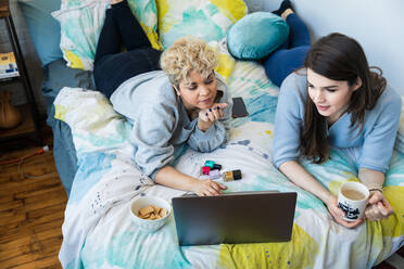 Hochformatige Ansicht von Freunden, die einen Laptop benutzen, während sie zu Hause auf dem Bett liegen - CAVF78053