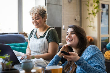 Happy young woman watching TV while friend using laptop at home - CAVF78054