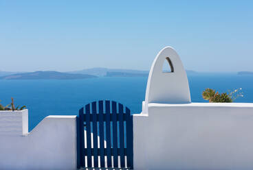 Fassade aus Ziegelsteinzaun oder kleiner weißer Mauer mit einem blauen Tor zum Ein- und Ausfahren und einem weißen Schornstein ohne Rauch mit dem blauen Ägäischen Meer im Hintergrund auf der idyllischen Insel Santorin in Griechenland - CAVF78087