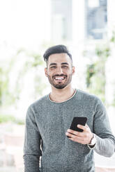Porträt eines lächelnden Geschäftsmannes, der ein Mobiltelefon hält, während er im Büro steht - MASF17555