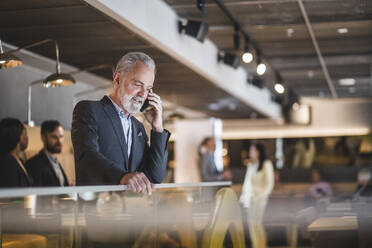 Älterer männlicher Unternehmer, der mit seinem Smartphone telefoniert, während er an einem Geländer im Büro steht - MASF17585