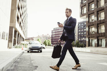 Male entrepreneur looking at phone while crossing street - MASF17625