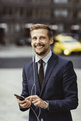 Porträt eines lächelnden Unternehmers, der im Freien stehend ein Smartphone über In-Ear-Kopfhörer benutzt - MASF17634
