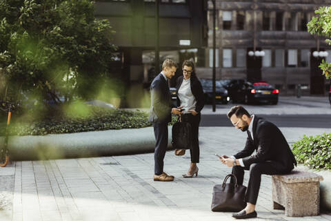 Geschäftsmann zeigt einer Kollegin sein Smartphone, während ein männlicher Kollege im Freien im Internet surft, lizenzfreies Stockfoto