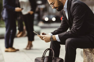 Businessman using smart phone while sitting on seat outdoors - MASF17641