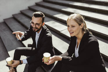 Smiling businessman pointing while sitting with female coworker on staircase - MASF17646