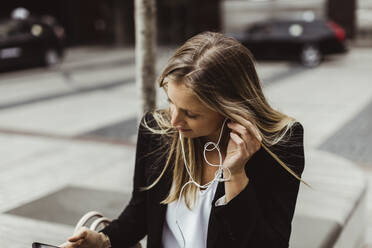 Geschäftsfrau mit In-Ear-Kopfhörern, die im Freien sitzt - MASF17662