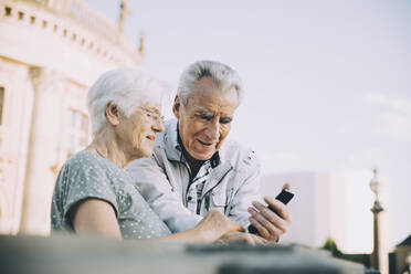 Senior male and female tourists using smart phone while standing in city against sky - MASF17668