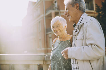 Lächelndes älteres Paar mit Arm um Spaziergang in der Stadt an einem sonnigen Tag - MASF17671