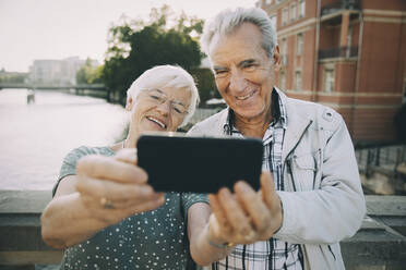 Lächelndes älteres Paar, das ein Selfie mit dem Handy macht, während es an einem Geländer in der Stadt steht - MASF17672